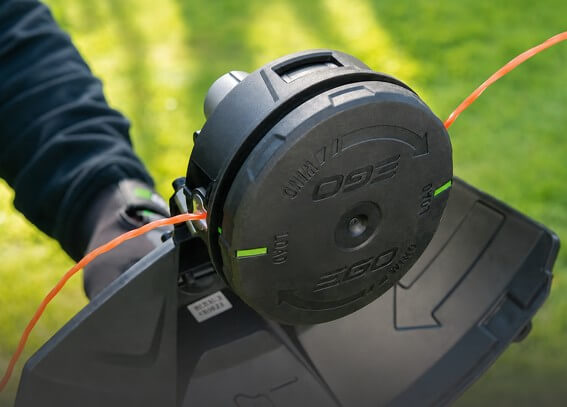 Close-up of a string trimmer head with orange line extended, mounted on a lawn tool with green grass in the background.